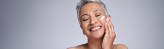 A woman with short gray hair applies moisturizer to her face with a smile. 