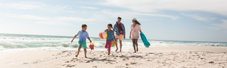 A mother, father, daughter, and son walk along a sandy beach with towels, a beach ball, and a pail and shovel for a fun day in the sun.