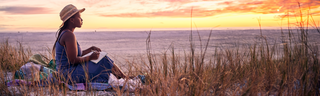 A young woman wearing a straw hat is relaxing with a book on a grassy beach, watching a colorful sunset