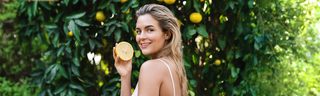 A blond woman in her 30s with shoulder length hair is wearing a whit tank top. She stands in front of a citrus tree with dark green leaves holding half of a grapefruit and smiling.