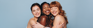Three diverse young women with beautiful skin smile against a blue background.