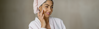 A smiling woman in a robe applies moisturizer as part of her healthy skin habits skincare routine.