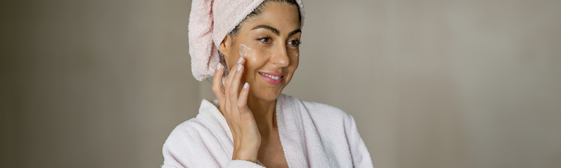 A smiling woman in a robe applies moisturizer as part of her healthy skin habits skincare routine.