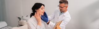 A woman in her 40s with dark hair looks at the results of a cosmetic procedure in the mirror as her doctor, a man in a white coat with glasses, examines her face.