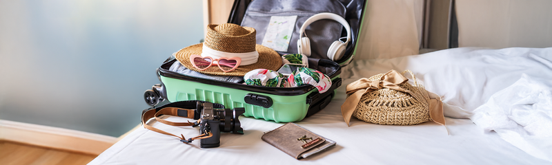 A bright green suitcase packed with colorful travel essentials like sunglasses, head phones, a camera, and a sun hat. 