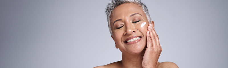 A woman with short gray hair applies moisturizer to her face with a smile. 