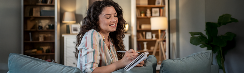 A smiling woman with long, dark curly hair sits on a green couch writing in a journal. 