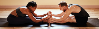 A happy couple wearing black athletic clothing, a man and woman are stretching together on black yoga mats in a sunny room.