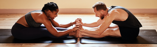 A happy couple wearing black athletic clothing, a man and woman are stretching together on black yoga mats in a sunny room. 