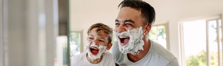 A man and his young son practice shaving in a sunny bathroom with shaving cream all over their smiling faces.