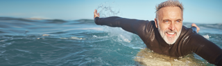 A smiling man with gray hair paddles on his surfboard in the ocean on a sunny day.