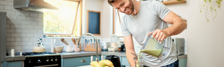 A smiling man makes a smoothie in his sunny kitchen while listening to music on headphones.