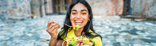 A smiling young woman in a yellow sweater eats a colorful salad on a sunny patio.