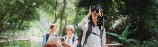 A group of women walk through the green woods with binoculars and a birdwatching guide book.. 