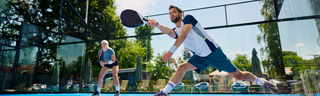 A man and woman in athletic clothing play pickleball on a sunny day on an outdoor court. The man raises his paddle to volley the ball. 