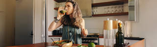 A young woman with long brown hair and a green shirt is drinking a green smoothie in a sunny kitchen. 