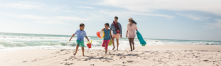 A mother, father, daughter, and son walk along a sandy beach with towels, a beach ball, and a pail and shovel for a fun day in the sun. 