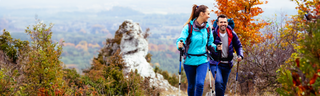 A smiling man and woman hike through a mountainous area with a beautiful landscape of red, yellow, and green autumn leaves behind them.