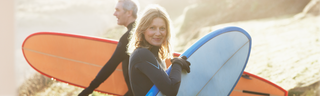 A smiling man and woman in their 50s, wearing wetsuits, carry their surfboards along a sunny beach on a cool fall day. 