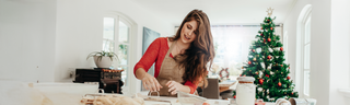 A woman with long brown hair wears a red shirt and brows apron. She is making cookies in front of a Christmas tree. 