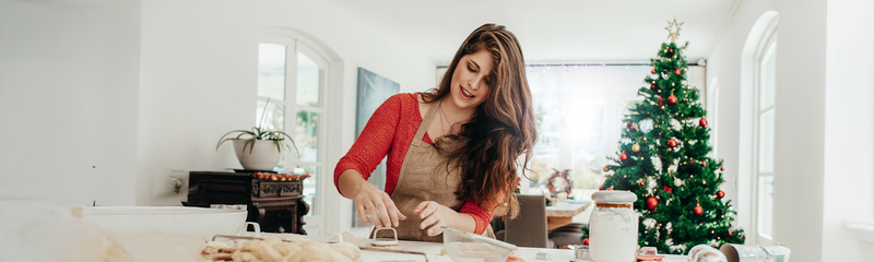 A woman with long brown hair wears a red shirt and brows apron. She is making cookies in front of a Christmas tree. 