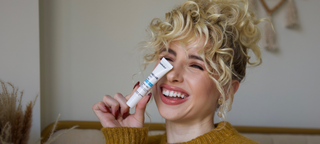 A young woman with blond, curly hair smiles as she prepares to apply arnica bruise cream to her face.