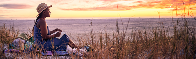 A young woman wearing a straw hat is relaxing with a book on a grassy beach, watching a colorful sunset