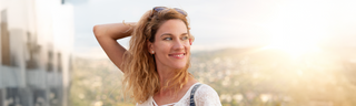 A woman in her 30s with blond curly hair stands in front of the sunset, smiling. 