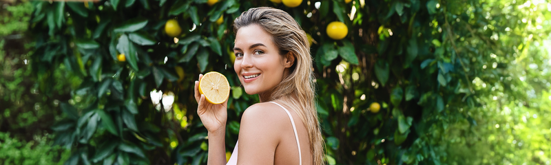 A blond woman in her 30s with shoulder length hair is wearing a whit tank top. She stands in front of a citrus tree with dark green leaves holding half of a grapefruit and smiling. 