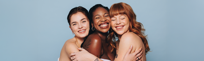 Three diverse young women with beautiful skin smile against a blue background. 