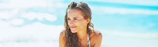 A caucasian woman in her 30s with clear skin and long brown hair relaxes on the beach. Blue water in the background. 