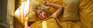 A blonde woman in her 30s sleeps in her bed with yellow sheets as the morning sun streams through the window. 