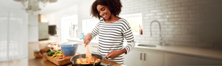A woman is stirring a pan of healthy vegetables on a stovetop in a bright kitchen.