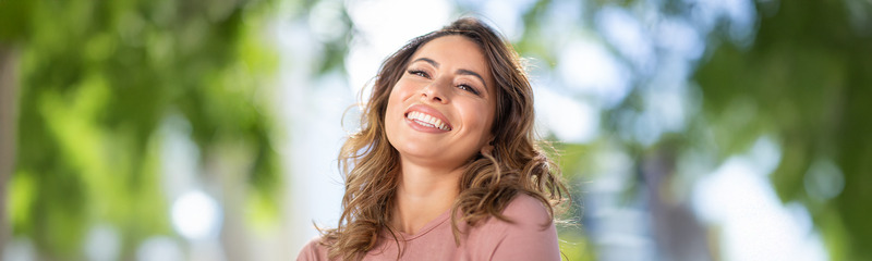 A smiling woman in her 40s shows off her glowing, youthful complexion and healthy hair. She has shoulder-length brown hair and wears a pink t-shirt, standing outside on a sunny day. 