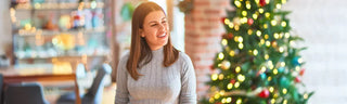 A woman with shoulder-length brown hair is wearing a gray sweater and smiling in front of her Christmas tree. 