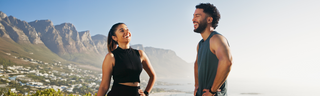 A man and woman of Latin descent are standing on the beach with cliffs in the background, wearing workout gear. They are smiling, and the sky is bright blue. 