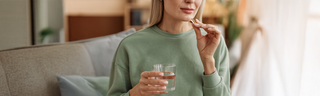 A caucasian woman in her late 30s with straight blond hair takes her fish oil supplements with a glass of water. She is wearing a green sweatshirt and sitting on a couch. 