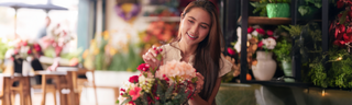 A women of Asian descent wears a pink shirt as she arranges flowers in a sunny room. She has long brown hair and is smiling brightly. 