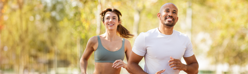 A caucasian woman with brown hair and an African American man with a beard in their 30s are running in the park. She wears a green sports bra and he wears a white t-shirt.  