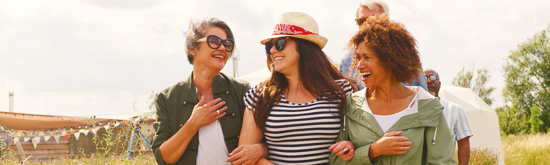 Three middle-aged women of different ethnicities walk arm-in-arm in the sunshine on an early, cool spring day. 
