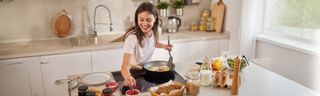 A woman with shoulder-length brown hair is making a healthy meal in her kitchen. She is smiling and wearing a whit t-shirt.