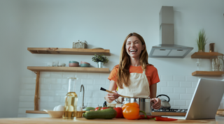 A smiling young blond woman in an organe t-shirt and white apron is making soup in a big metal pot in a sunny kitchen.